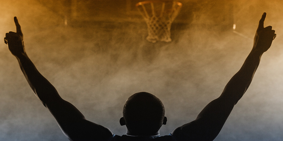 A basketball player with arms up is silhouetted against a smoky scene with a basketball hoop