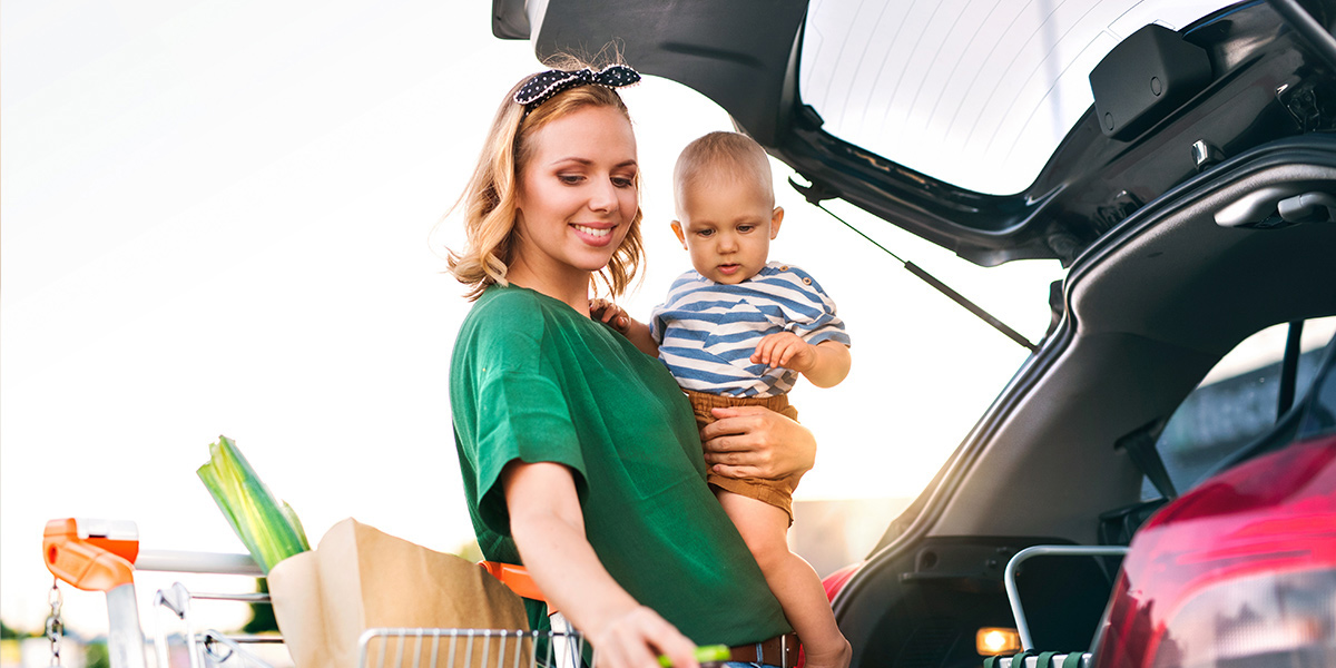 A single mother holding a baby loads groceries into the back of her car.