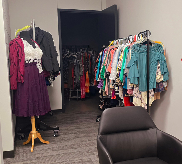 image displays a a photograph taken of the Clothing Closet existing inside the Student Resource and Advocacy Center. On the left, a mannequin is shown wearing a knee length dress in a purple color, decorated with white lace. To the right, a rack displays a selection of different clothing items available. 